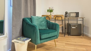 Un fauteuil turquoise avec un coussin, une petite table d’appoint en bois avec un livre, un réfrigérateur compact, un four à micro-ondes et une machine à café posés sur une table en métal.