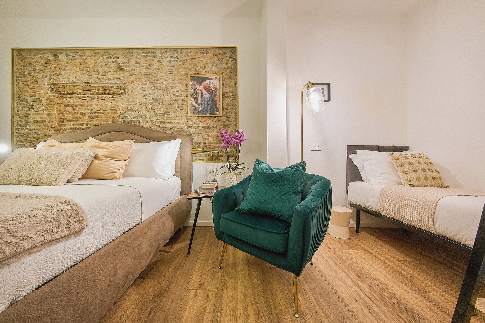 A hotel room with two beds, a green armchair, a stone wall, and a framed picture.