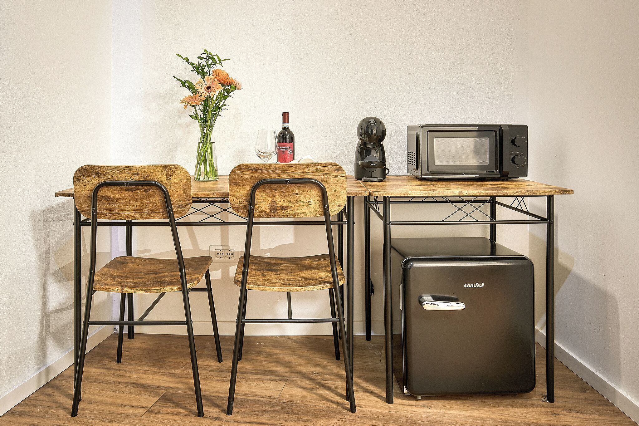 Uma kitchenette compacta com micro-ondas, máquina de café e mini-frigorífico. Há duas cadeiras de madeira e um vaso com flores na mesa.