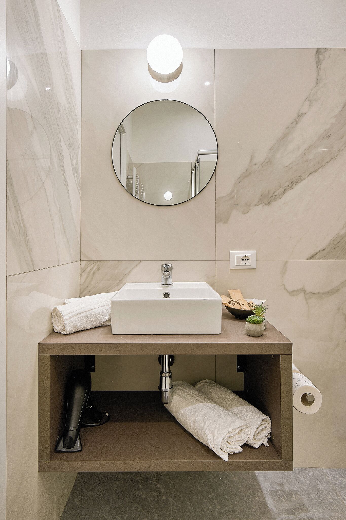 A bathroom with a white sink, marble walls, a round mirror, and a towel rack.