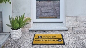 Une porte blanche avec un panneau de verre, un paillasson sur lequel est écrit « Arrivées » et « Départs », une plante en pot et un mur de pierre.
