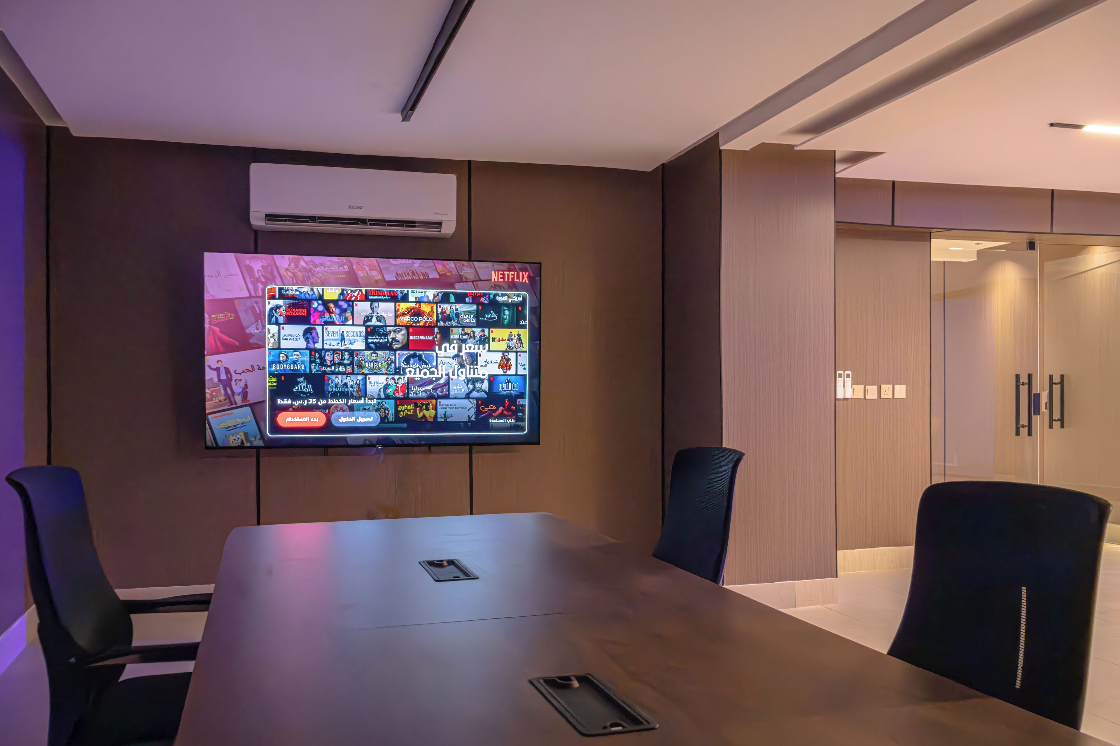Een vergaderruimte met een groot flatscreentelevisie waarop een Netflix interface te zien is, een lange tafel en zwarte stoelen.