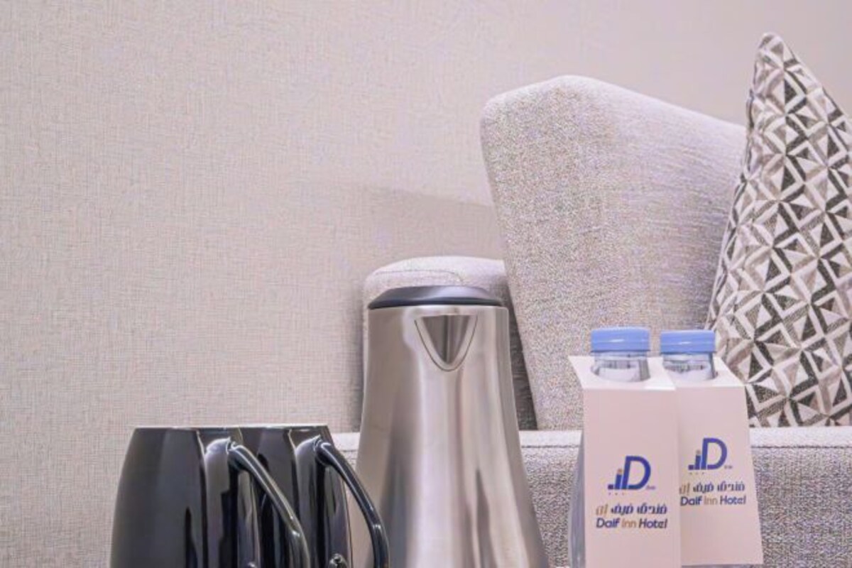A room with a sofa, a metallic kettle, a black bag, and a box of bottled water with the hotel logo.