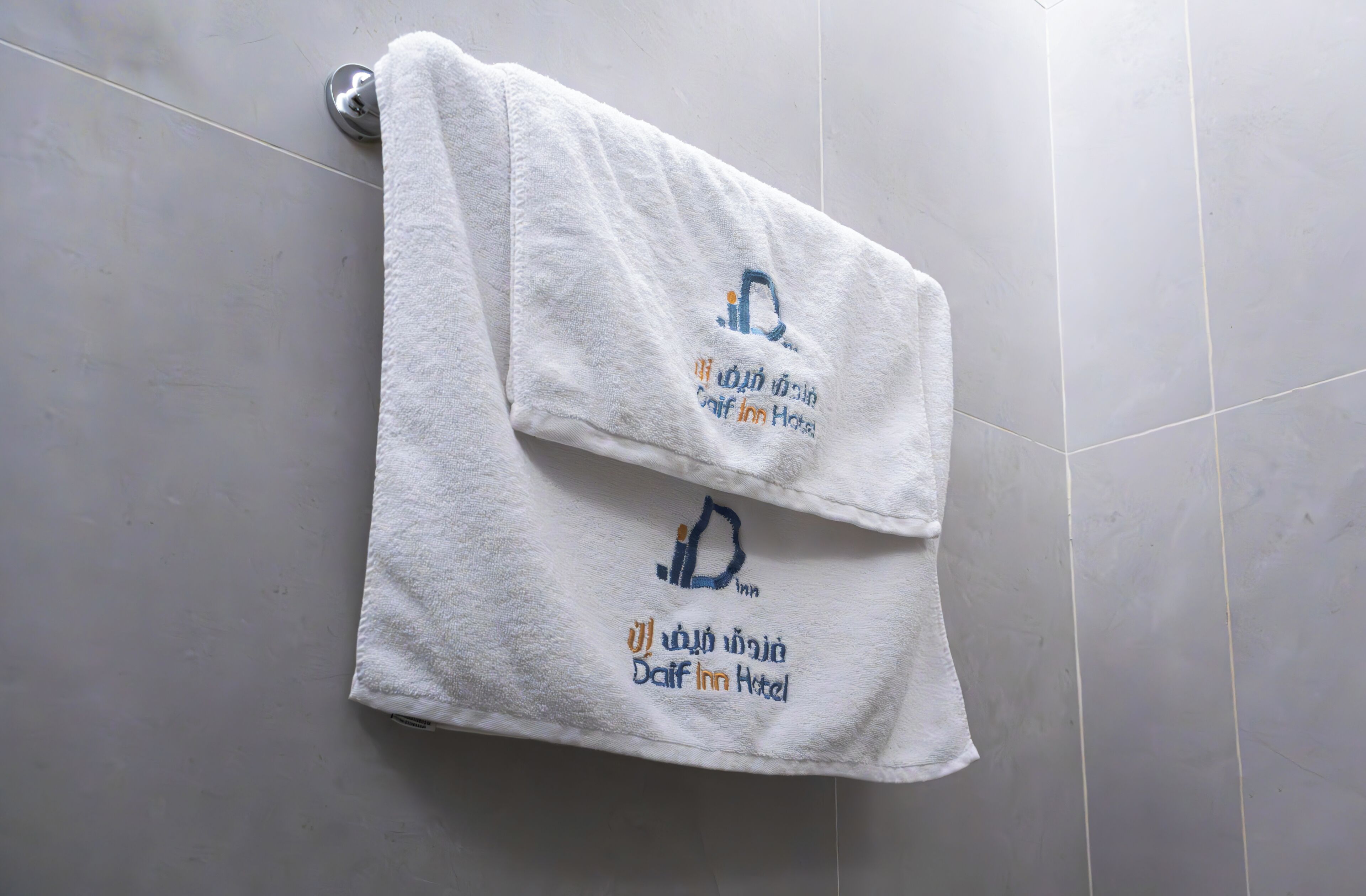 Une serviette blanche ornée du logo et du nom de l’hôtel Daif Inn est accrochée à un mur carrelé.