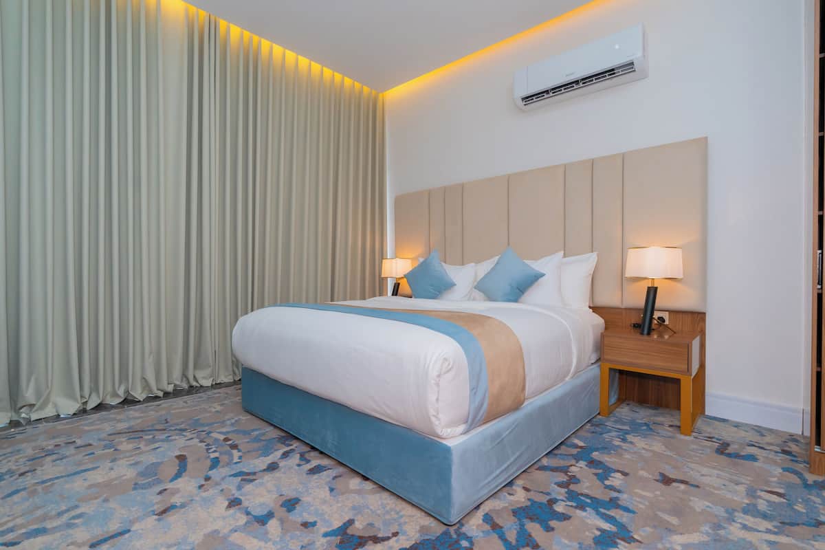 A hotel room with a large bed, two bedside tables with lamps, a wall-mounted air conditioner, and a patterned carpet.
