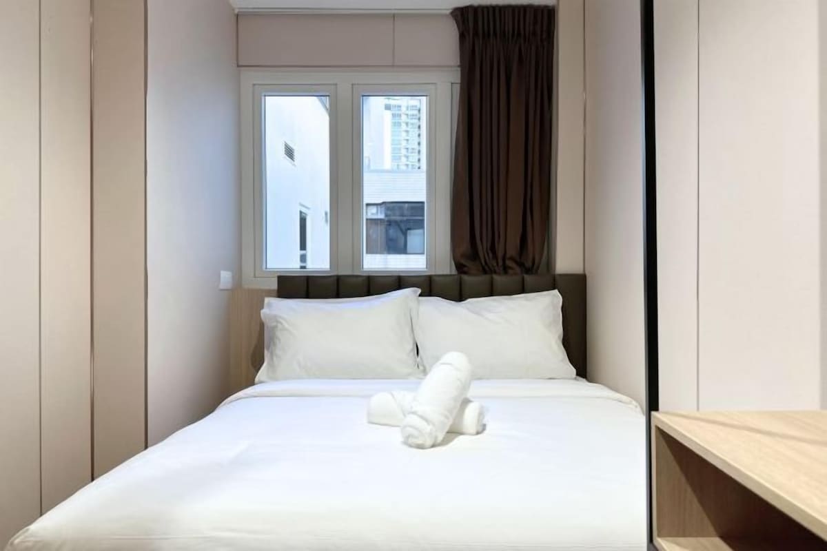 Una cama bien hecha con sábanas blancas y una toalla enrollada, una ventana con cortinas y vistas a edificios.