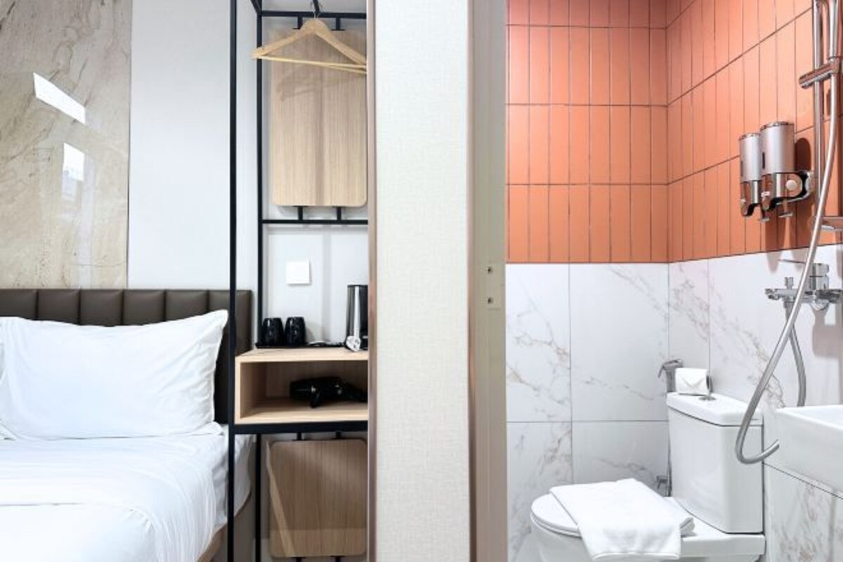 A modern hotel room with a bed, a wooden shelf with toiletries, a hanging coat, and a bathroom with a shower, toilet, and marble walls.