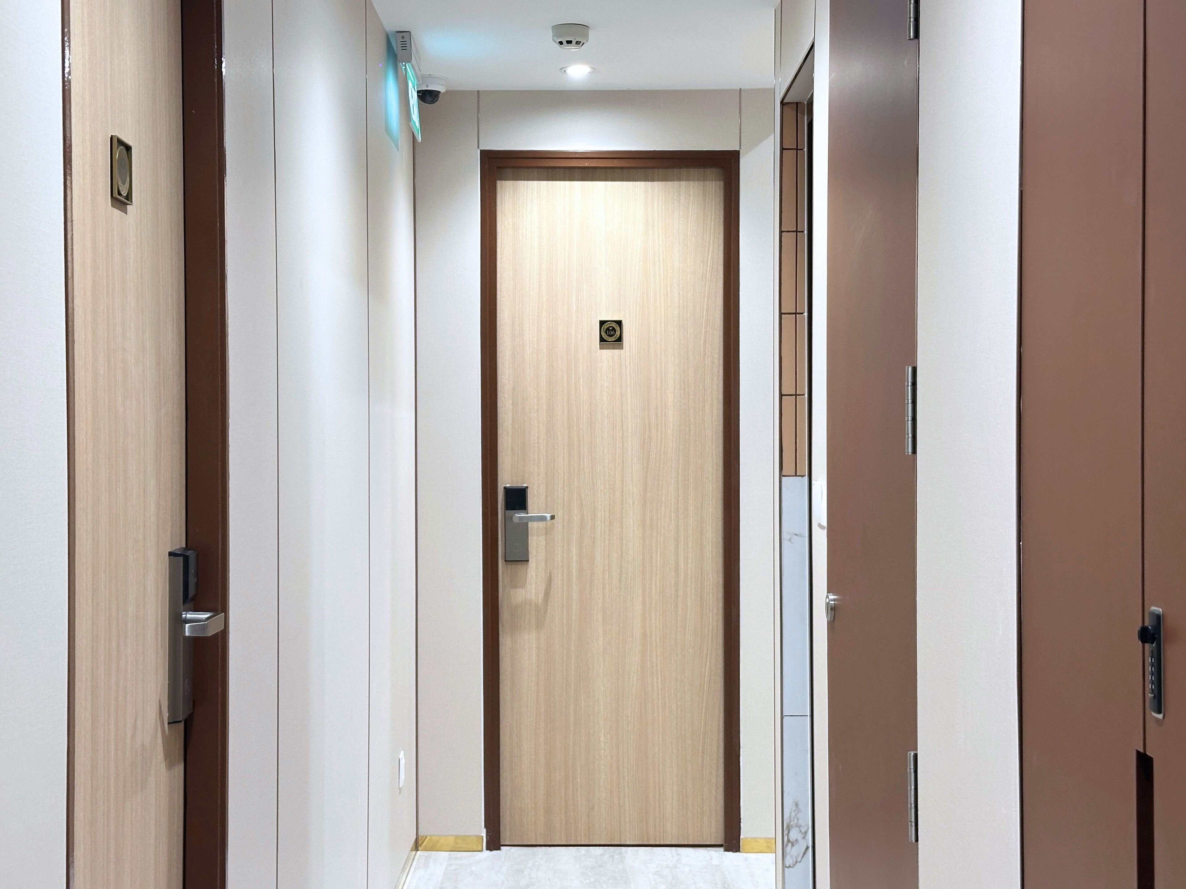 A narrow hallway with a wooden door at the end, a security camera, and a fire alarm on the wall.