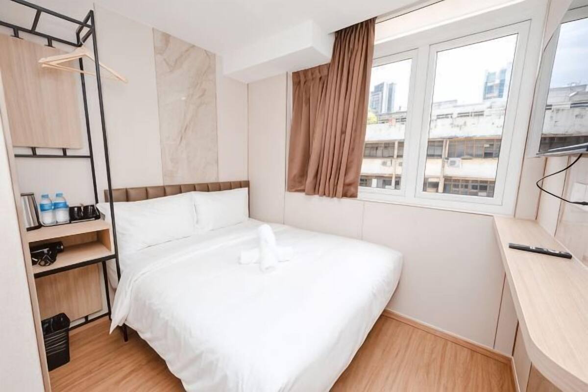 A hotel room with a bed, a desk, a window with curtains, a shelf with toiletries, and a hanging rack.