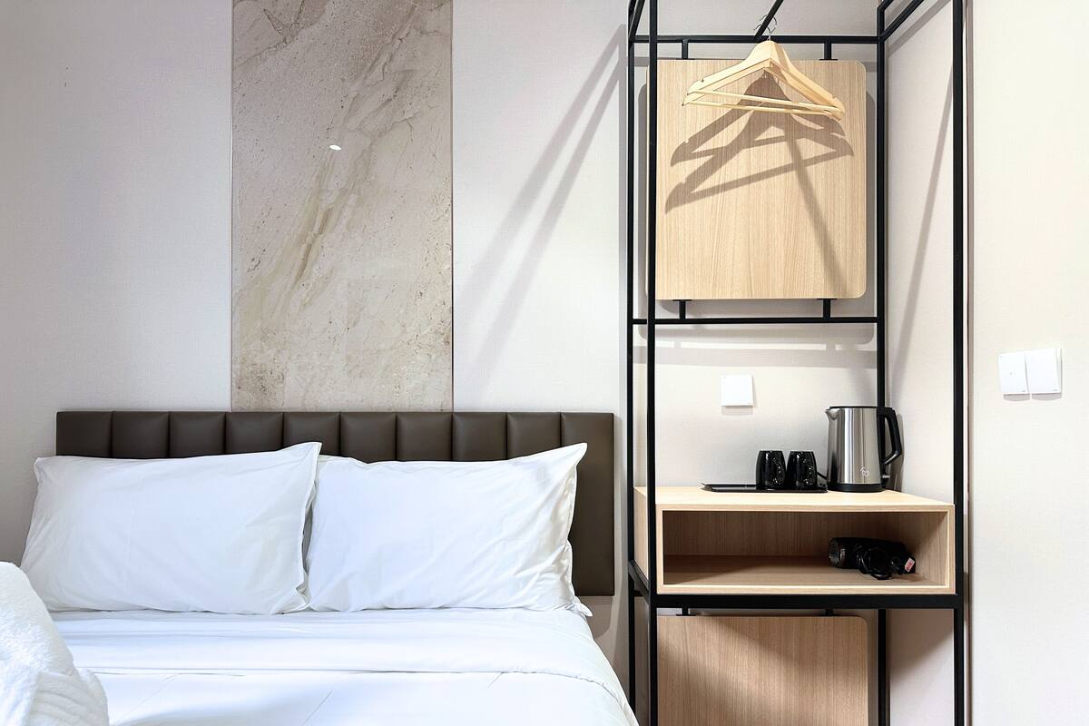 A hotel room with a bed, a wooden shelf with a kettle, and a hanging garment rack.