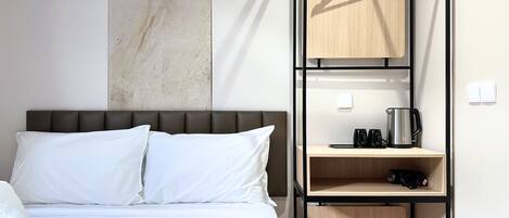 A hotel room with a bed, a wooden shelf with a kettle, and a hanging garment rack.