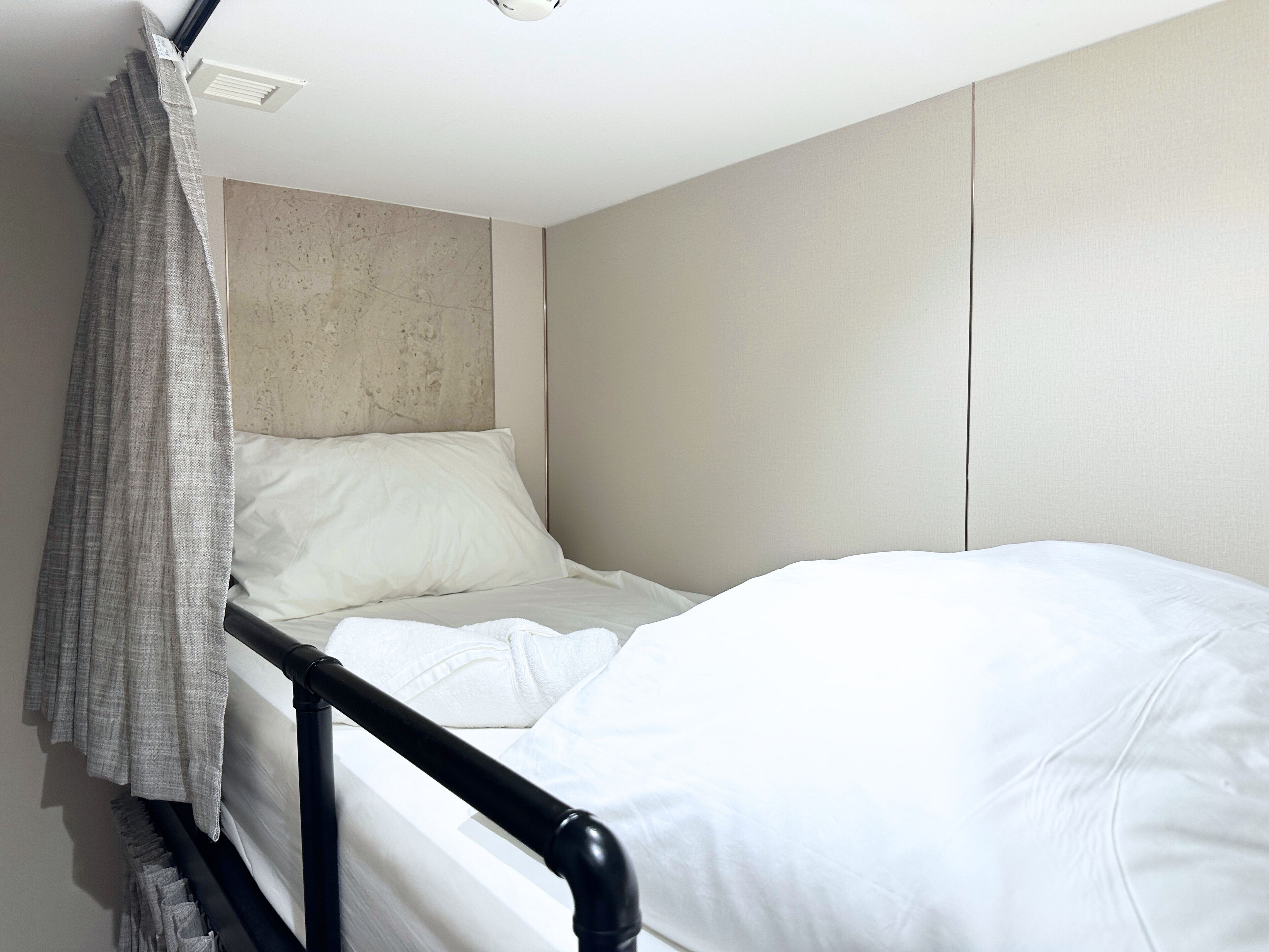 A single bed with white bedding and a grey curtain, a headboard with a textured finish, and a plain wall with a recessed light fixture.