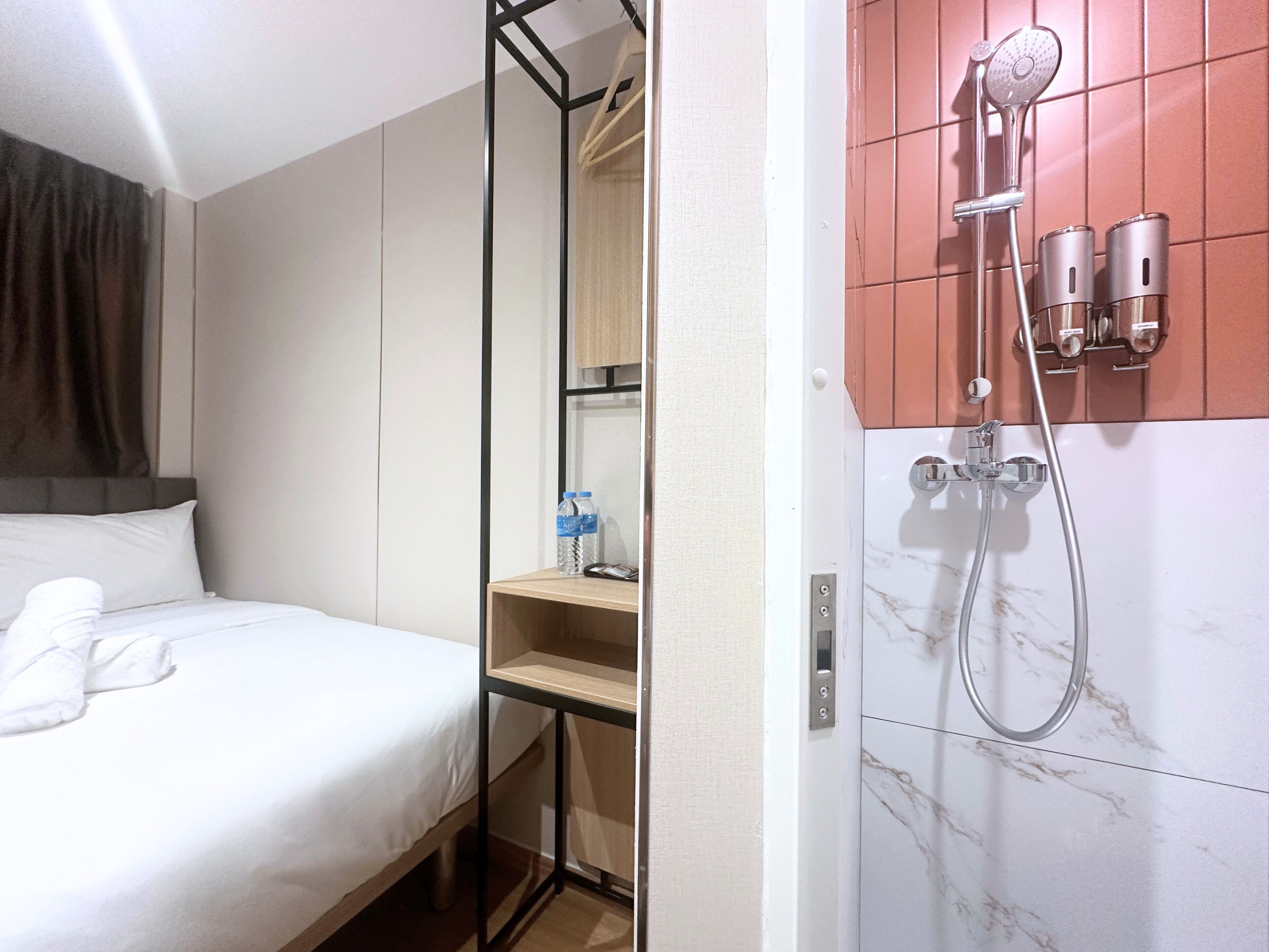 Camera d'albergo con un letto, una mensola in legno con articoli da toilette e un angolo doccia con soffione a mano e due flaconi.