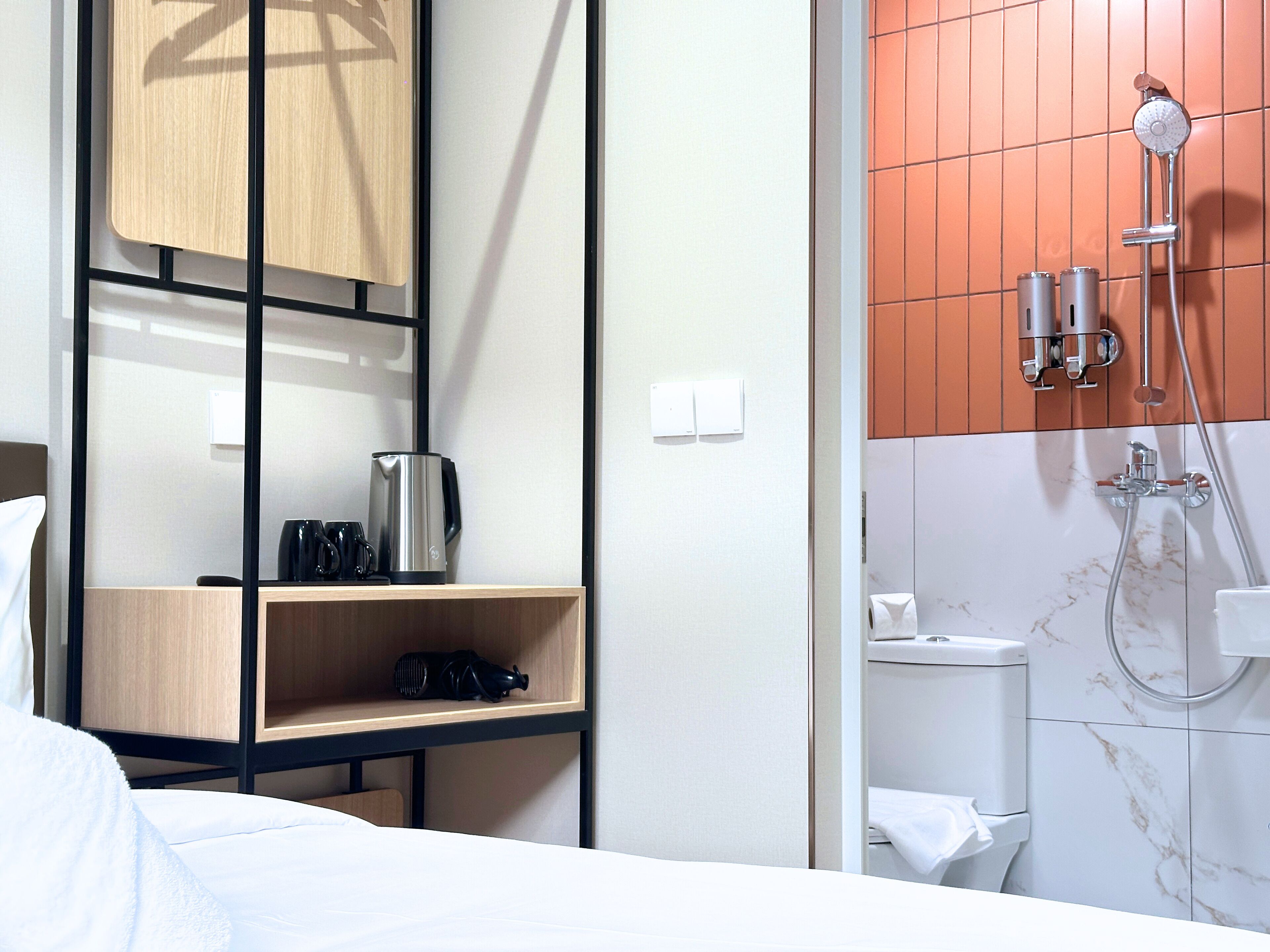 Habitación de hotel con cama, estante de madera con hervidor y tazas, ducha con cabezal de ducha manual y un inodoro.