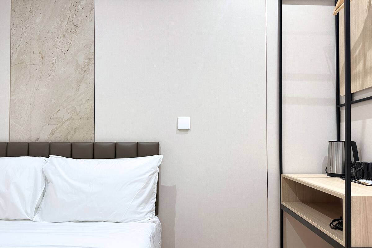A modern hotel room with a bed, a marble wall, a wooden shelf with a kettle, and a power outlet.