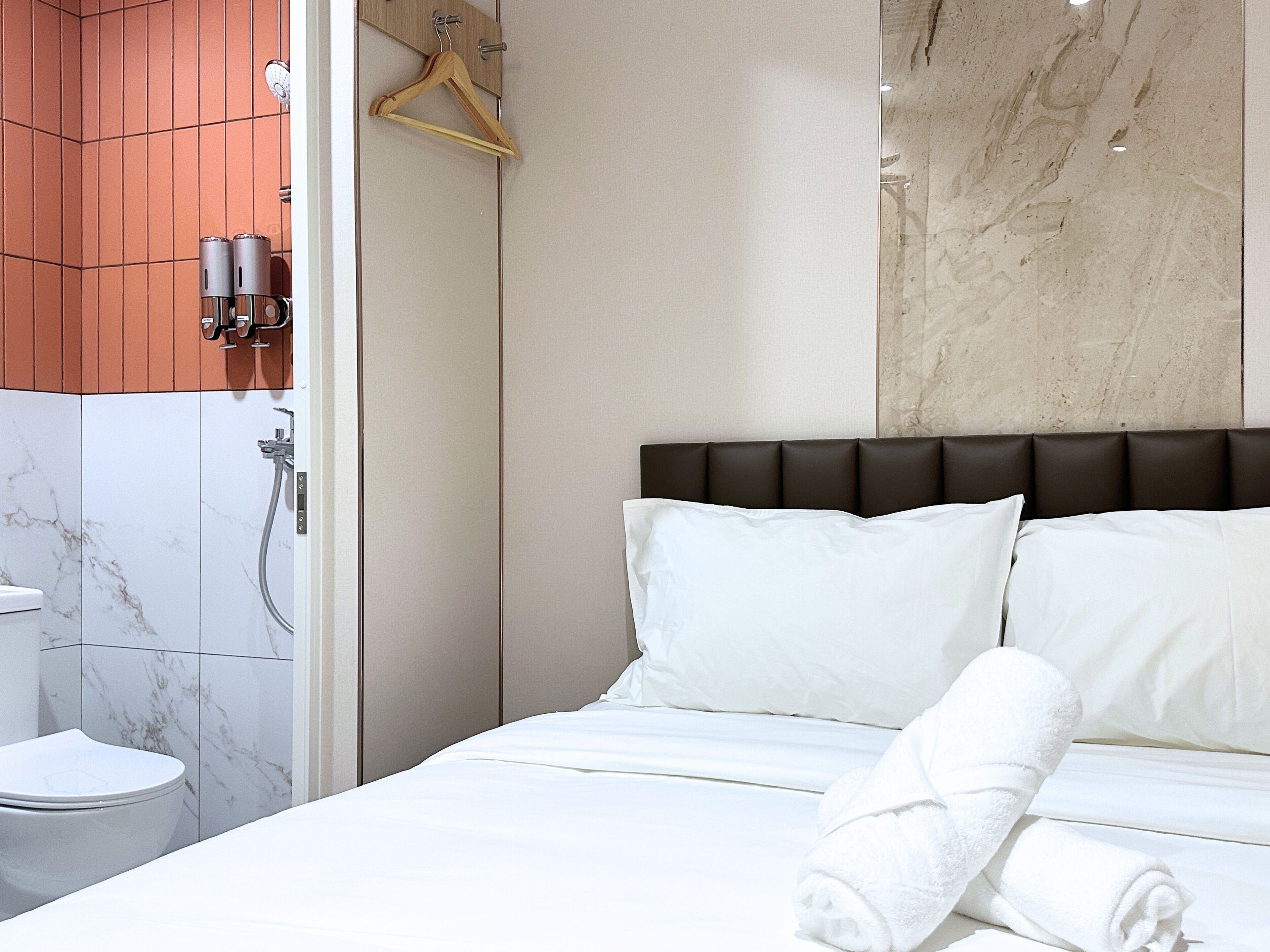 Une chambre d’hôtel comprenant un lit, du linge de lit blanc, un cintre en bois, des toilettes et un espace douche.
