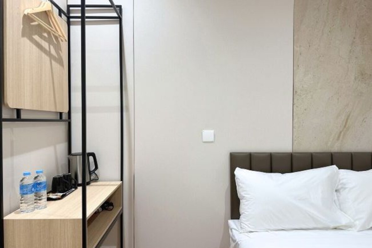 A hotel room with a bed, a wooden shelf with a kettle, and a water bottle.