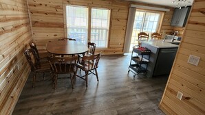A wooden cabin interior with a dining table and chairs, a kitchen area with a sink and cabinets, and large windows allowing natural light.