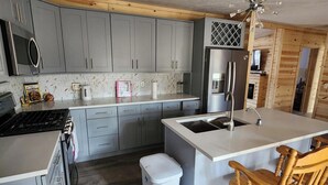 A modern kitchen with grey cabinets, a stainless steel refrigerator, a built-in island with a sink, and a wooden chair.