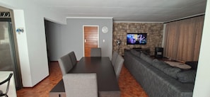 A modern living room with a long dining table, grey sofas, a flat-screen TV mounted on a stone wall, and a wooden door.