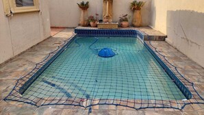 Una pequeña piscina exterior con una cubierta de malla azul, rodeada por una zona de terraza con baldosas.