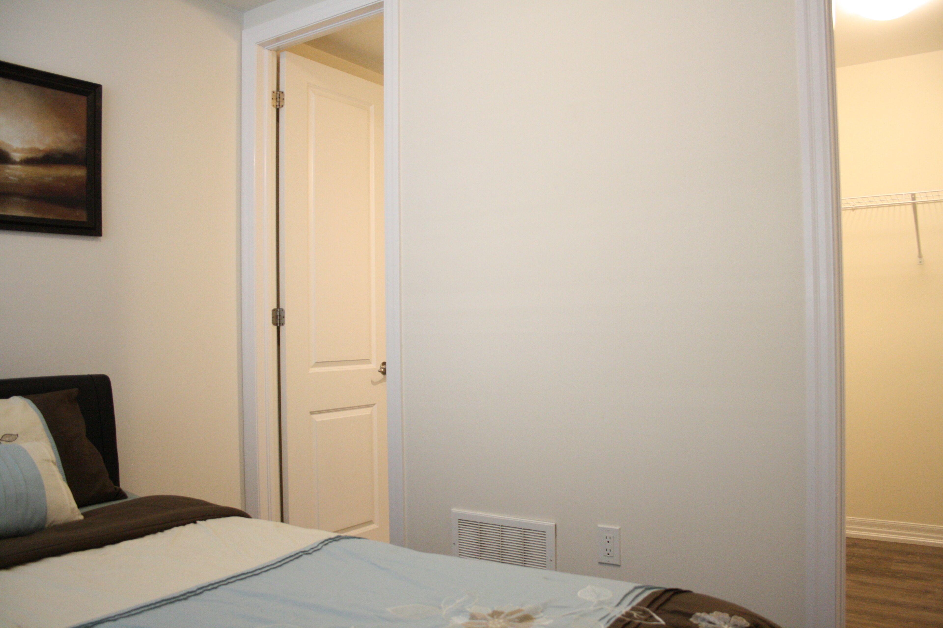 Een slaapkamer met een bed, een deur naar een andere kamer, een wandlamp en een ventilatie rooster.