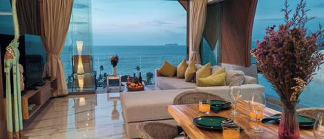 A modern hotel room with a large window offering a view of the sea, a cozy seating area with cushions, a dining table set with glasses and a vase of flowers.