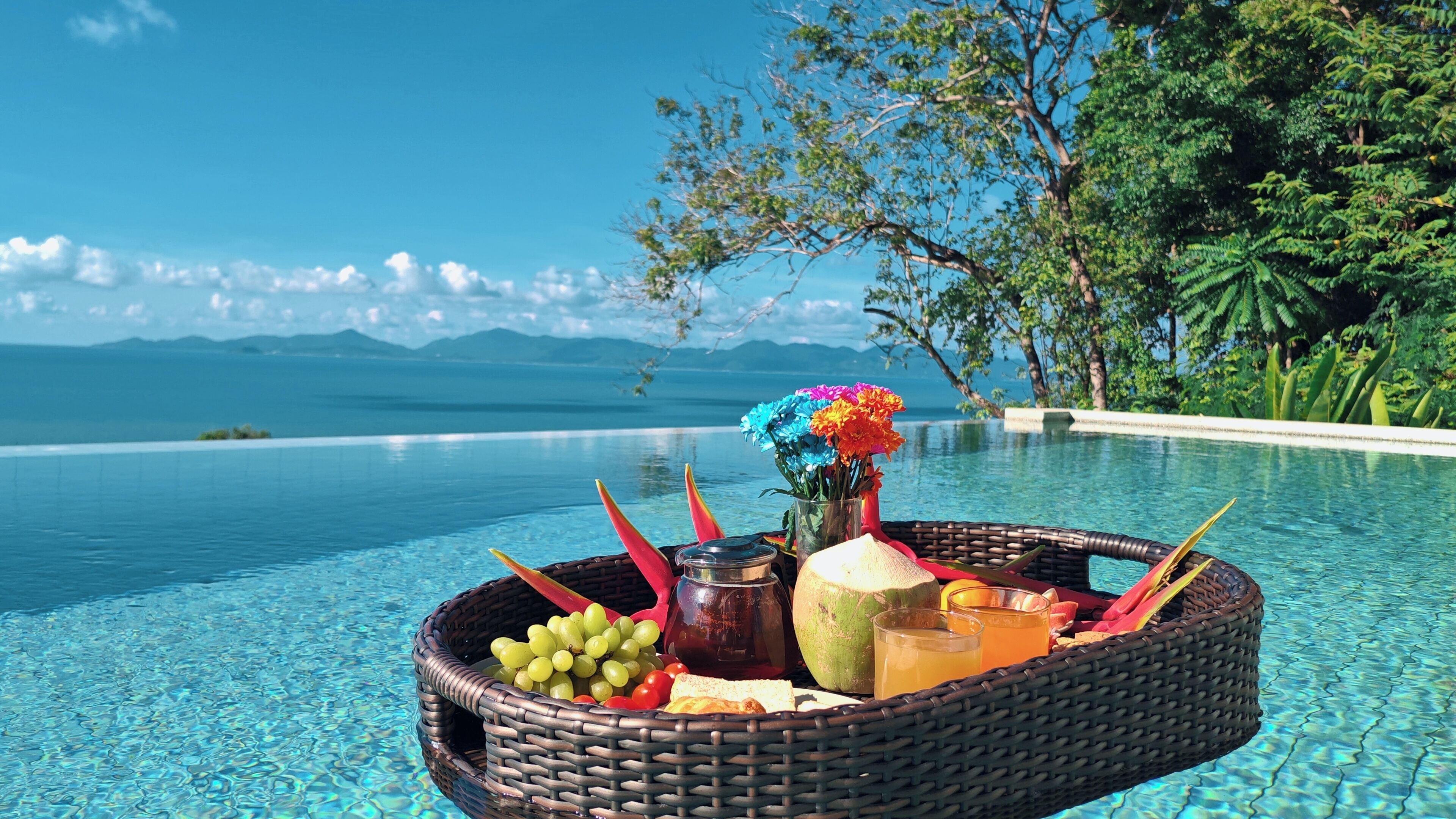 A wicker basket filled with fruits, a coconut, and drinks on a poolside with a clear view of the ocean and mountains in the background.