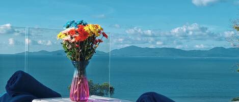 Vista despejada de un paisaje costero con montañas al fondo, una mesa de vidrio con un florero de flores coloridas y cojines azules en un banco.