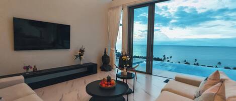 A modern living room with a large TV, comfortable seating, a small table with fruit, and a view of the ocean.