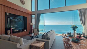 A modern living room with a large TV, comfortable seating, a dining area with a table set for two, and a stunning ocean view through large windows. - Santi Pura Villas (Koh Samui)