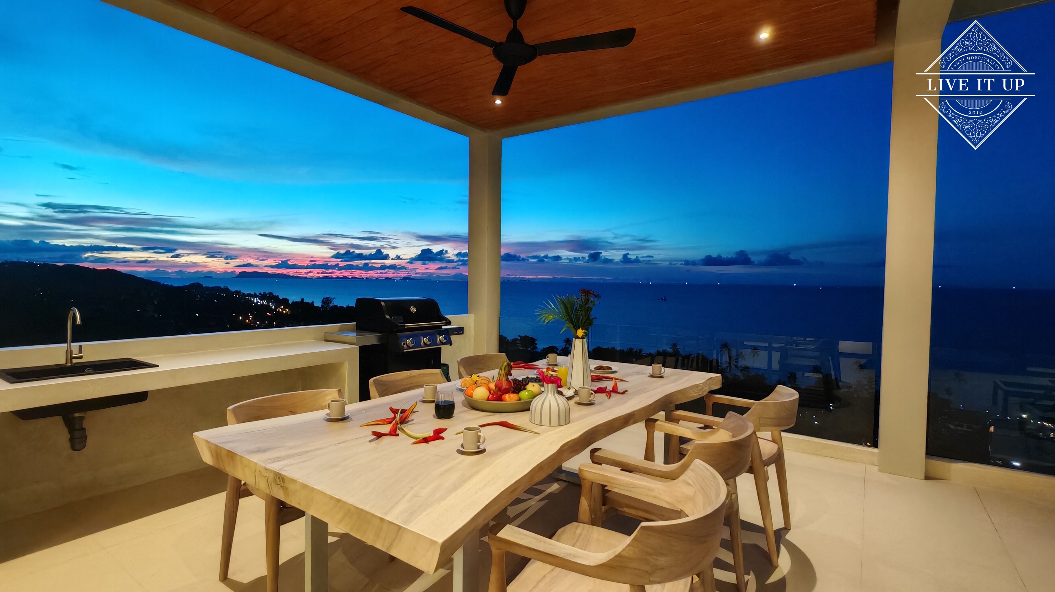 A dining area with a view of the ocean at dusk, a ceiling fan, and a grill.