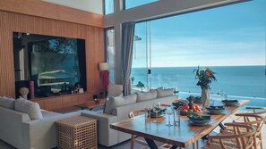 Un salon moderne doté d’un grand téléviseur, de sièges confortables, d’un coin repas avec une table dressée pour deux personnes et d’une vue sur l’océan.