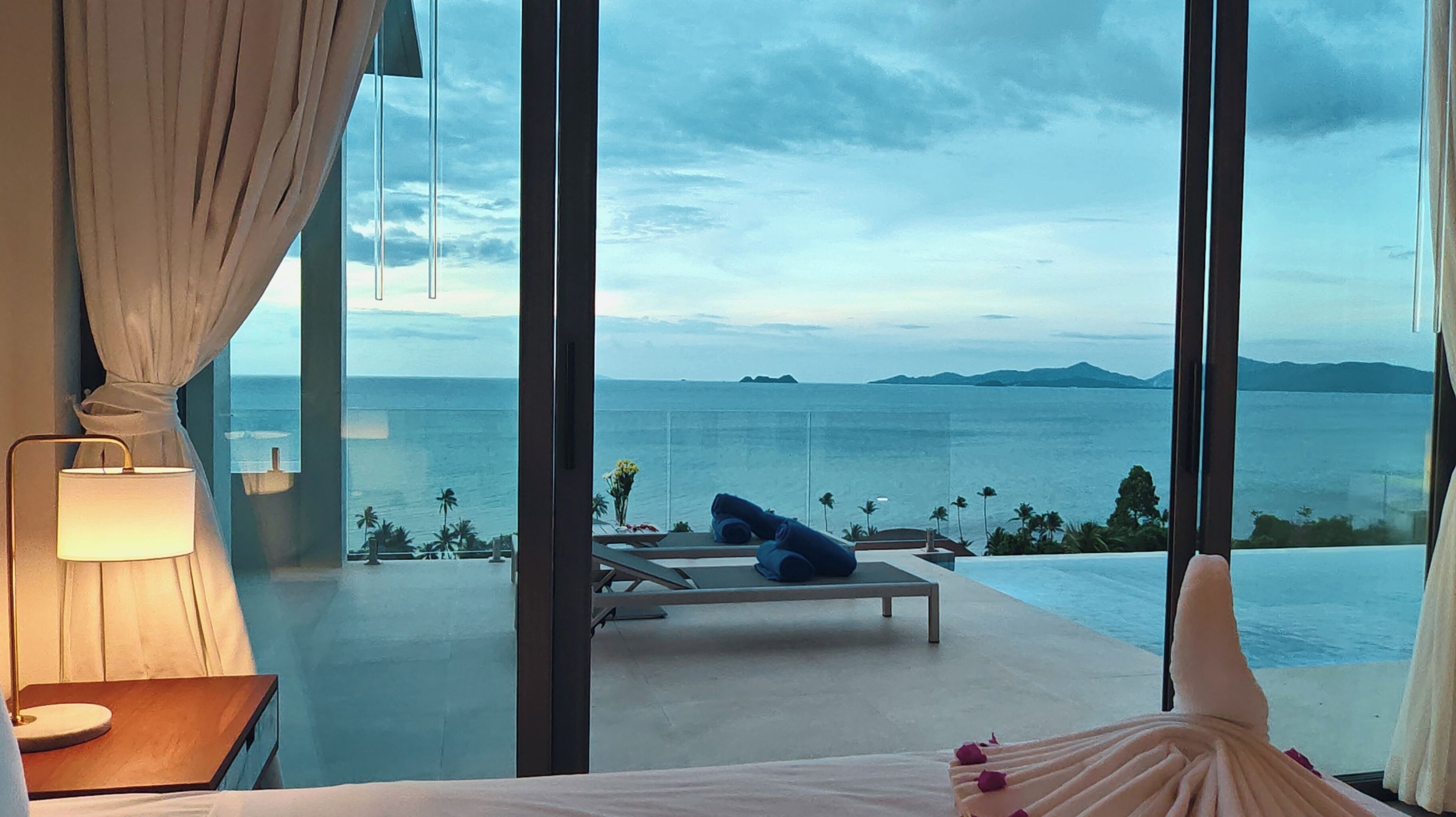 Un dormitorio con un amplio ventanal con vistas a una piscina y al mar. Hay dos sillones y una mesa con lámpara.