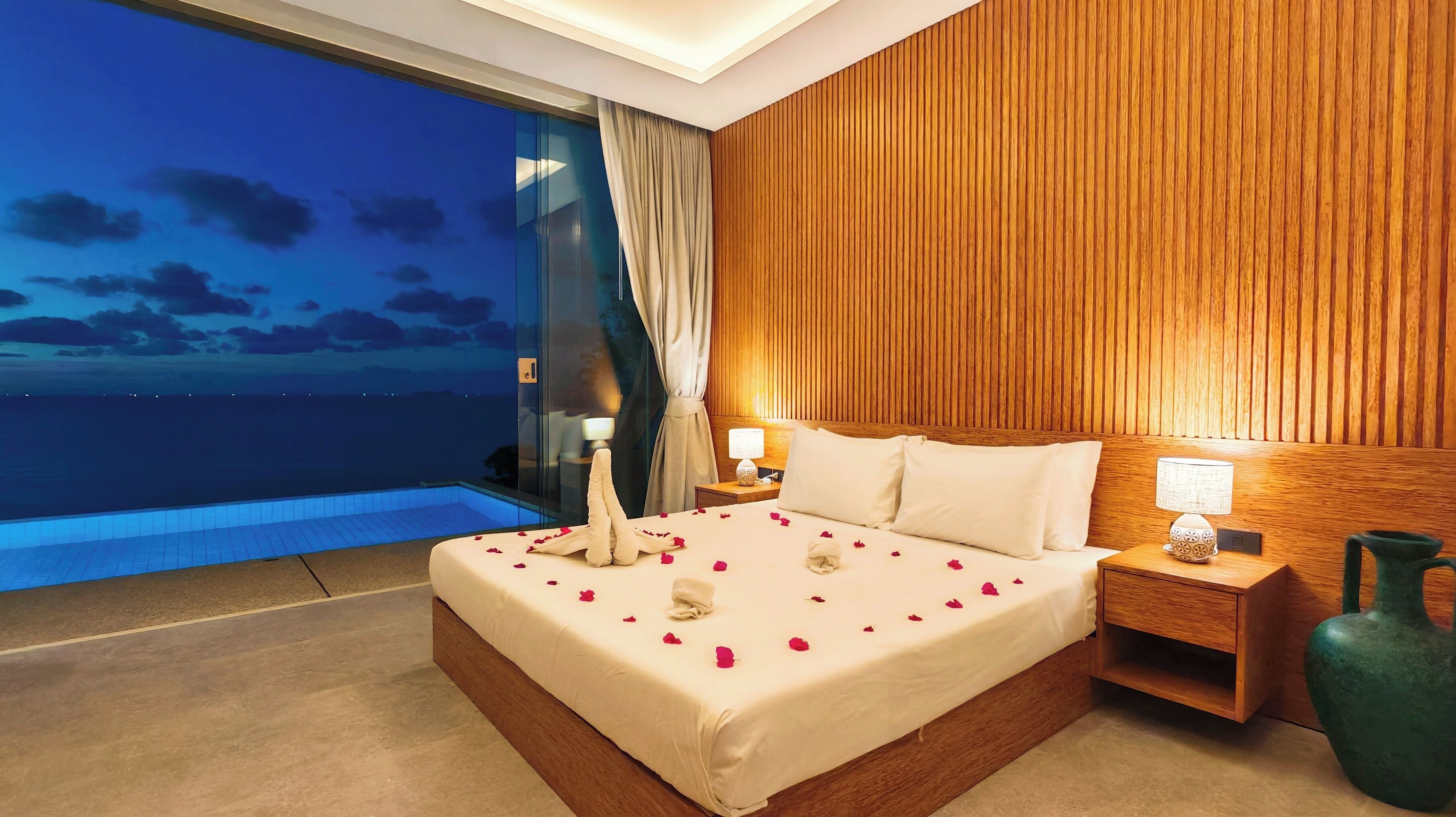 A hotel room with a large bed, a nightstand, a wooden panel wall, and a view of the sea.