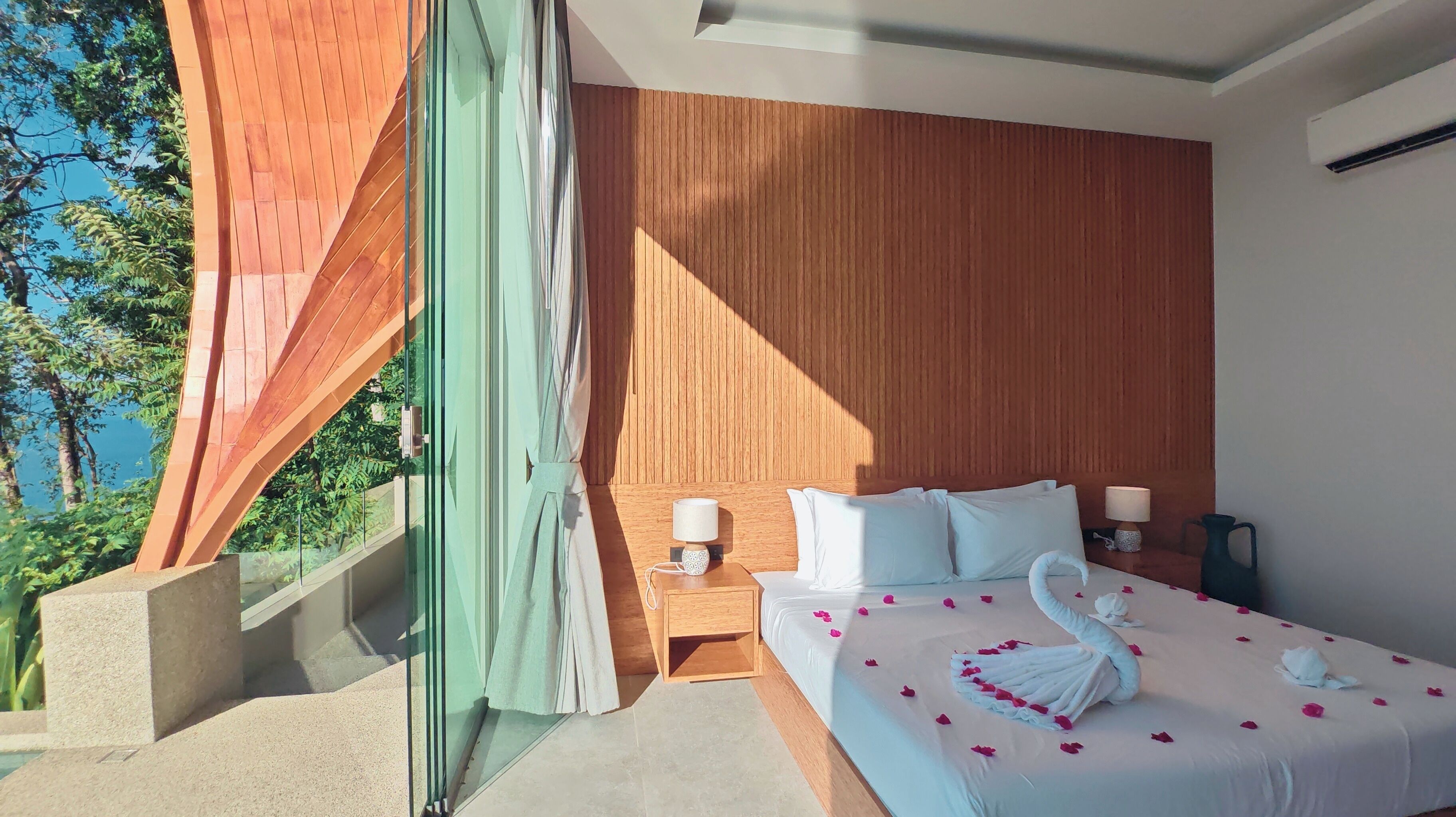 A modern hotel room with a large bed, a nightstand, and a view of greenery.