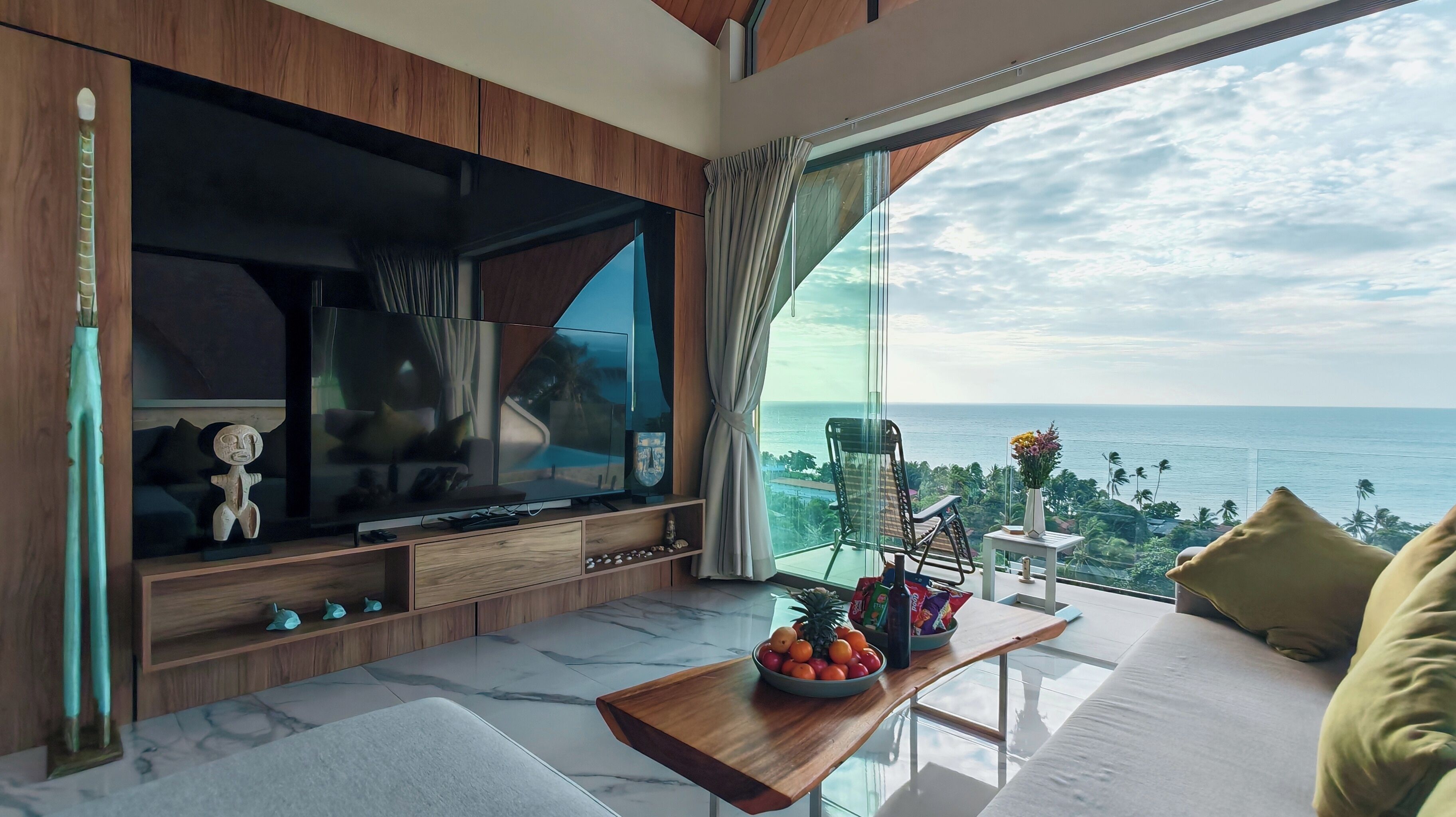 Una sala de estar moderna con un televisor de pantalla plana grande, muebles de madera, piso de baldosas de mármol, una mesa de centro de vidrio con fruta y vista al océano.