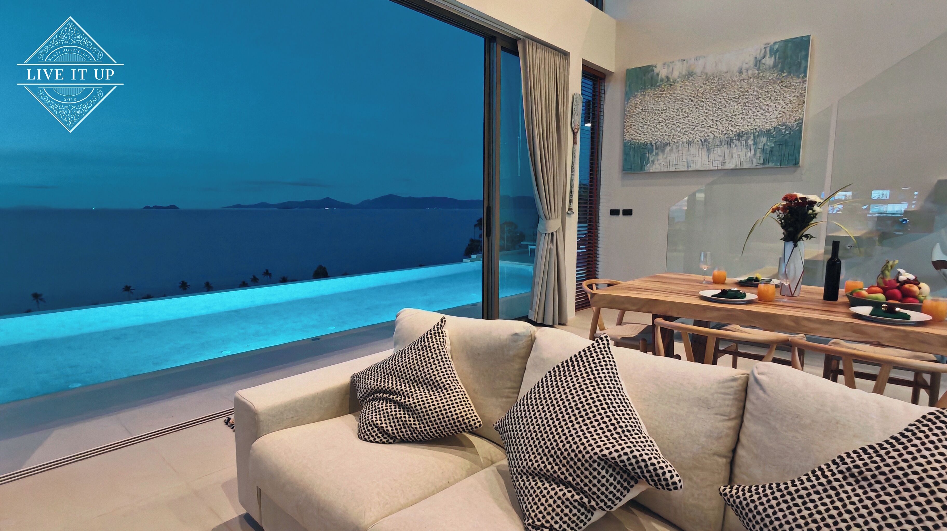Ein modernes Wohnzimmer mit großem Fenster mit Blick auf Pool und Meer. Es gibt eine Essecke mit Tisch für zwei Personen und eine Vase mit Blumen auf dem Tisch.