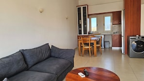 A compact living space with a grey sofa, a wooden dining table with chairs, a kitchen area with wooden cabinets, and a washing machine. - Cassiopeia 3-Bedroom House on beach with pool (Chania)
