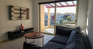 A modern living room with a sofa, a coffee table, and a TV mounted on the wall. There is a sliding glass door leading to a balcony with a view of buildings and greenery. - Cassiopeia 3-Bedroom House on beach with pool (Chania)