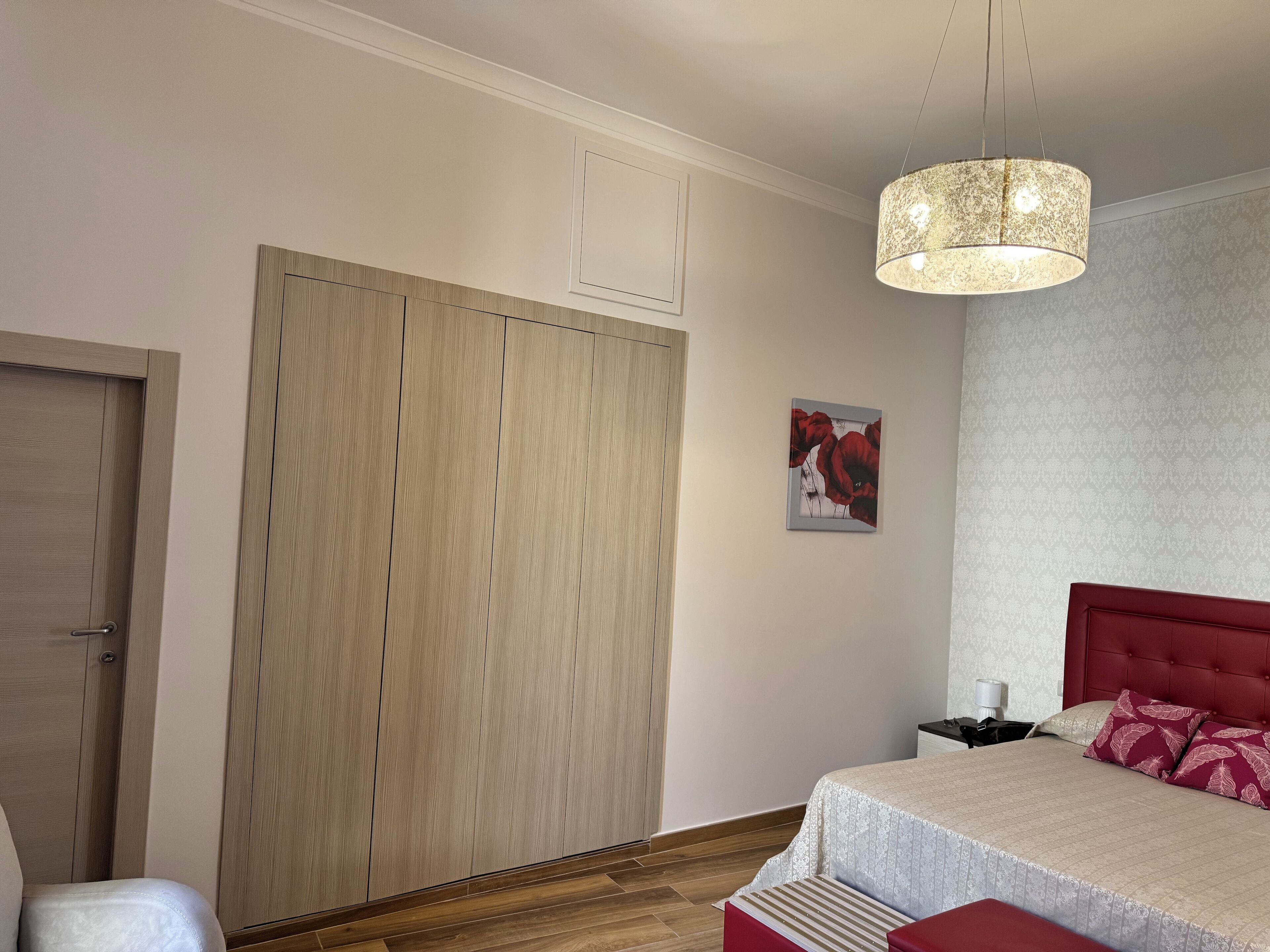 A bedroom with a large wardrobe, a bed with a red headboard, a hanging light fixture, and a framed picture on the wall.