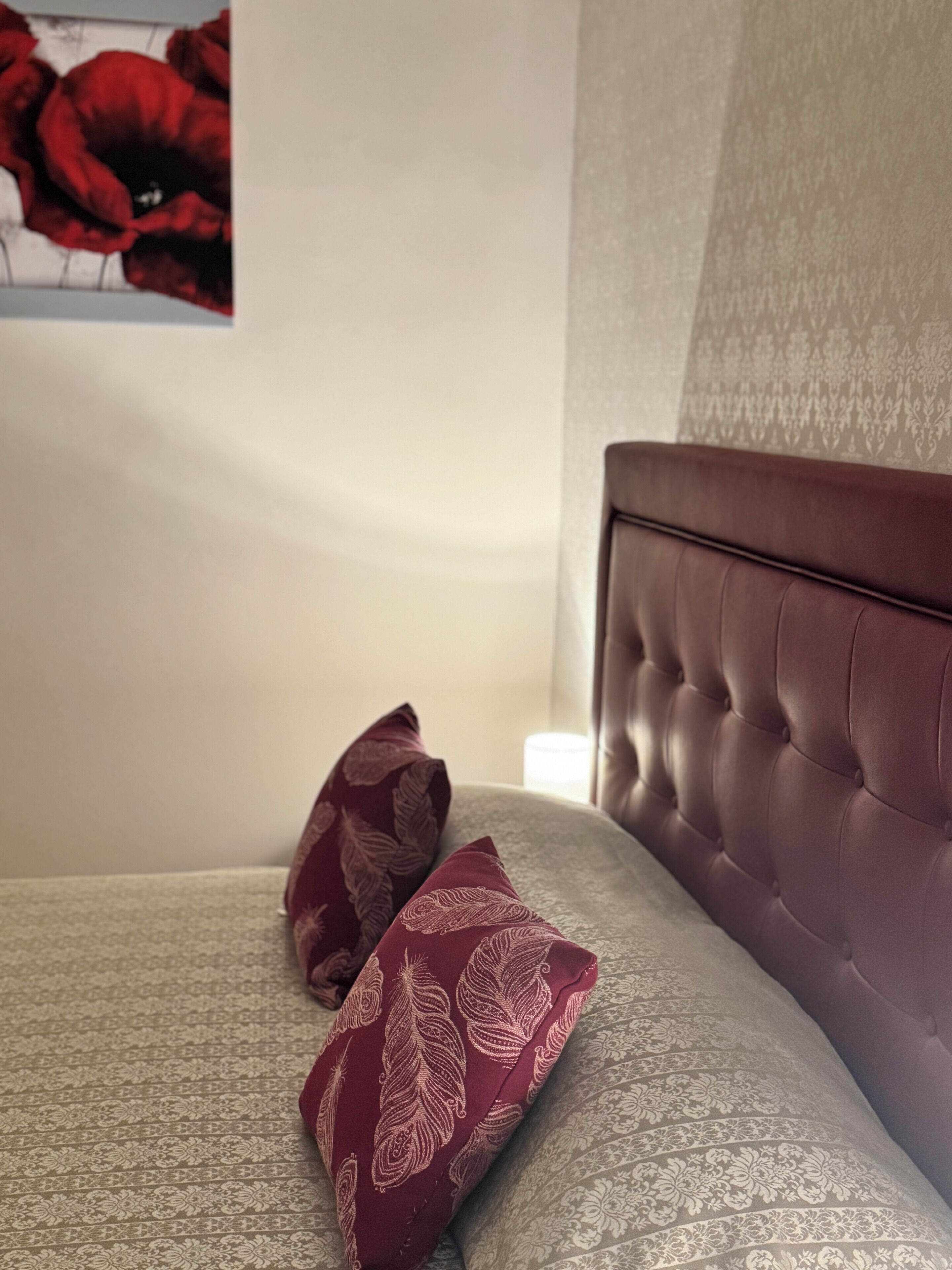 A neatly made bed with a tufted headboard, two patterned pillows, and a wall with a floral painting.