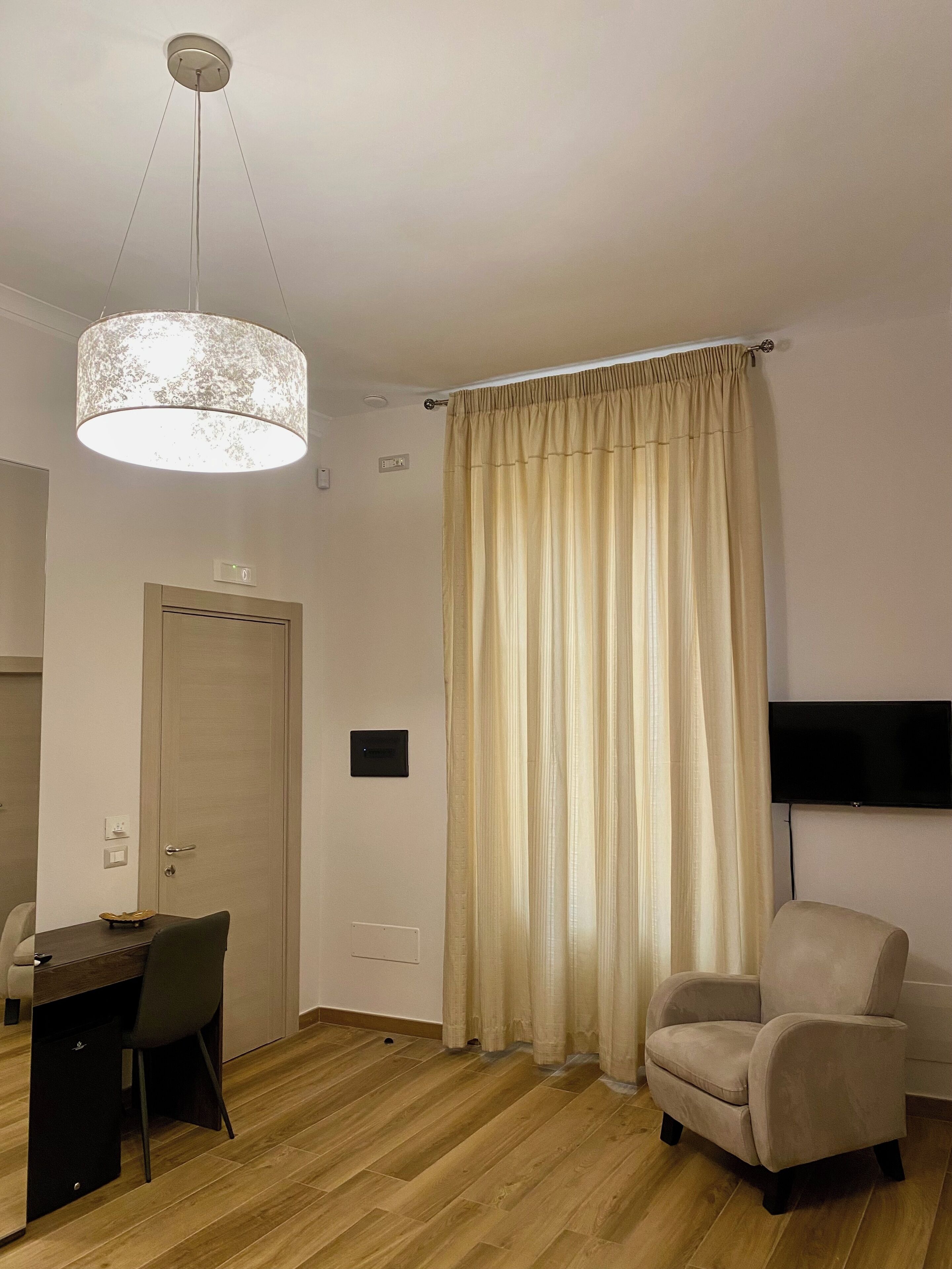 Ein modernes Wohnzimmer mit Holzboden, einem Esstisch mit Stühlen, einer Couch, einem an der Wand befestigten Fernseher und einem großen Fenster mit beigen Vorhängen.