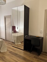 Un dormitorio con un armario espejo, una cama con ropa de cama estampada, un escritorio con una silla y una pequeña mesa con objetos encima.