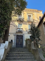 Un edificio con una entrada central, una escalera que lleva a ella y un árbol en primer plano.