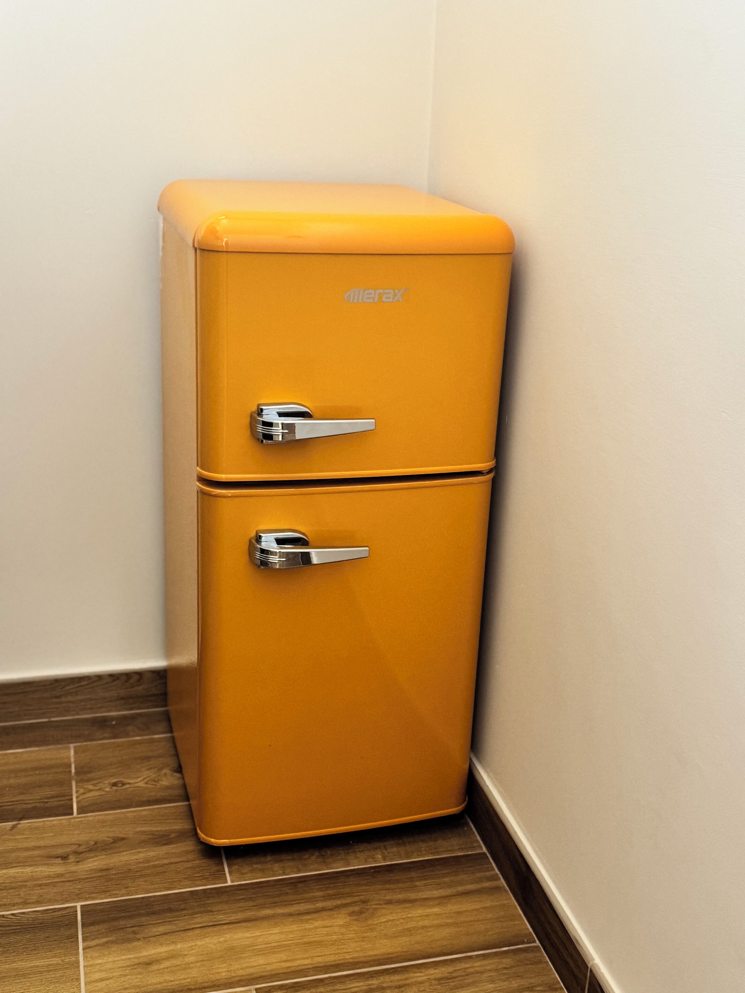 A compact, two-door, orange vintage refrigerator with a metallic handle, placed in a corner against a white wall and wooden floor.