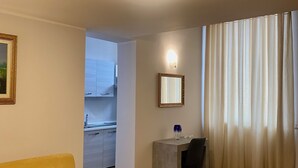 Un salon moderne avec un canapé jaune, un petit bureau doté d’un miroir et une vue sur l’extérieur à travers une fenêtre ornée de rideaux légers.