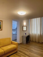 Una sala de estar moderna con un sofá amarillo, un escritorio pequeño con un espejo y vistas al exterior a través de una ventana con cortinas ligeras.