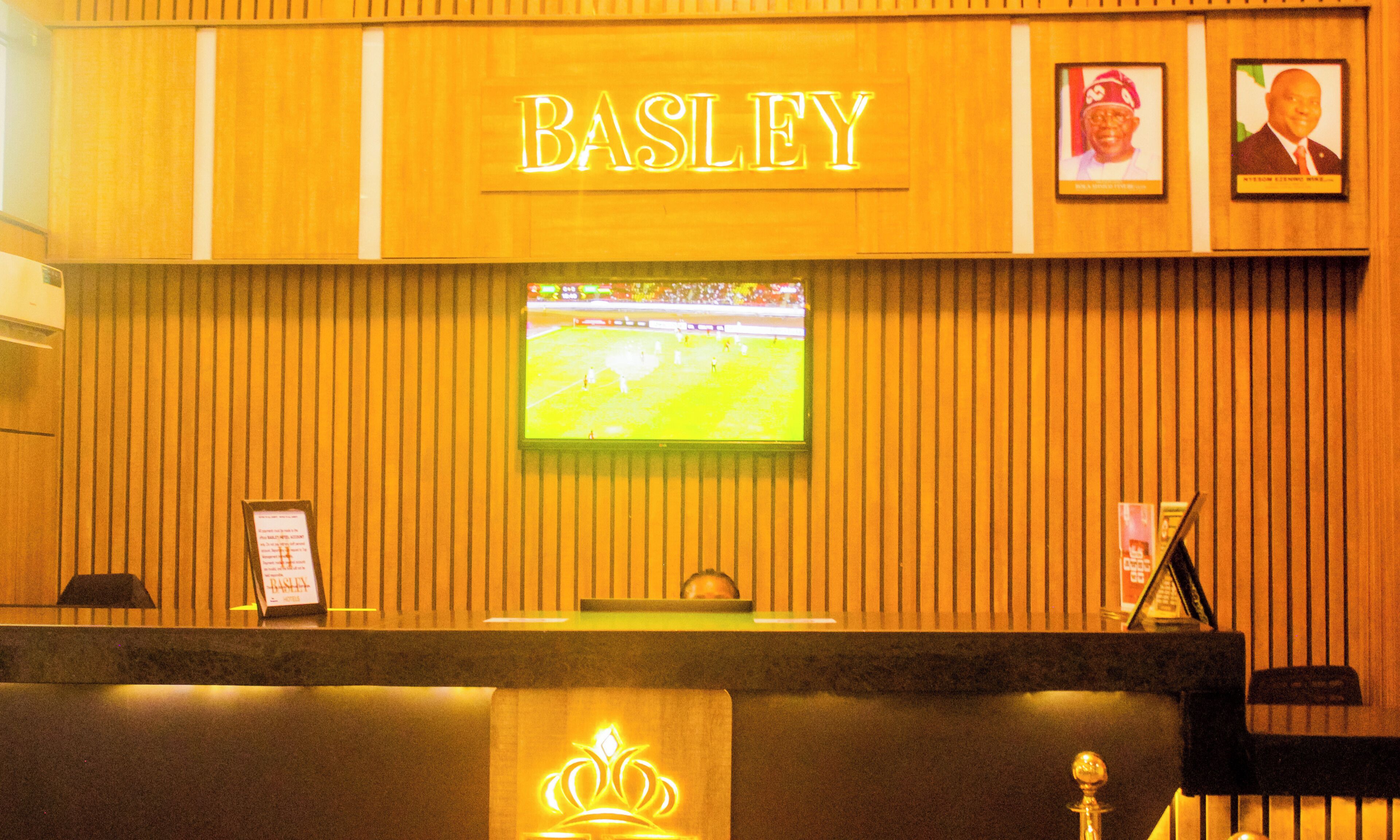 Ein Empfangstresen mit einem 'BASLEY'-Schild, ein Fernseher, der ein Sportereignis zeigt, und gerahmte Bilder an der Wand.