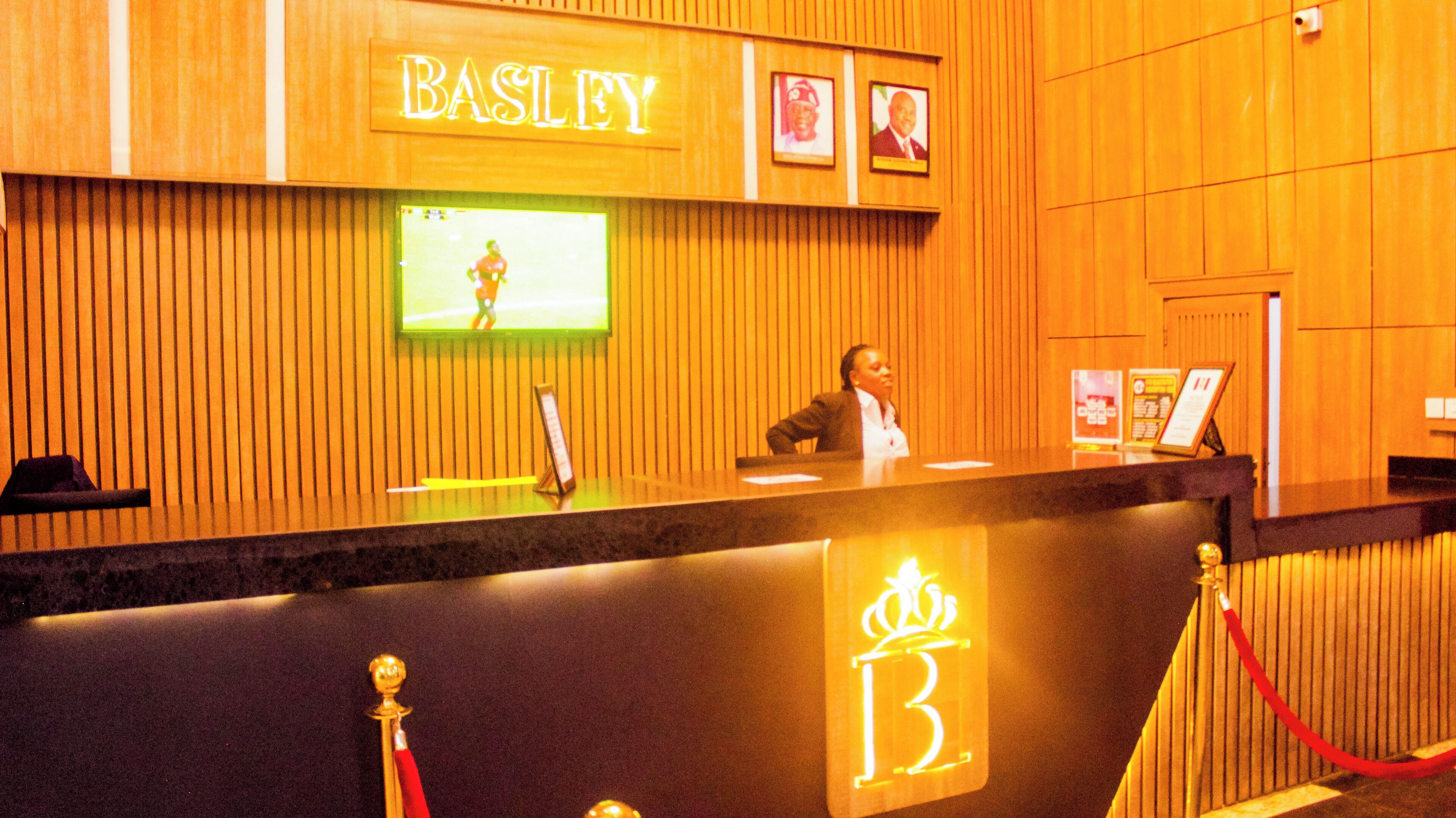 A hotel reception desk with a 'BASLEY' sign, a television screen, and framed pictures on the wall.